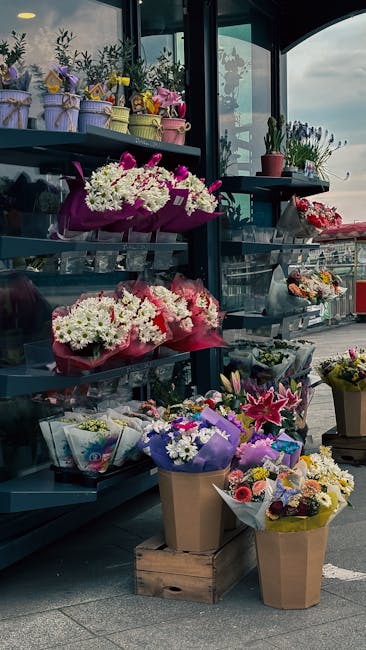 A display of various floral arrangements and bouquets at a flower stand. The arrangement includes vibrant white and pink chrysanthemums, lilies, and roses wrapped in purple, pink, and beige tissue pap