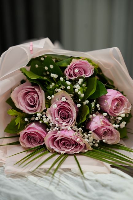 A bouquet of fresh, pale pink roses with tightly spiraled petals, accented by small white baby's breath flowers and lush green leaves. The flowers are arranged in a tightly clustered, rounded style an