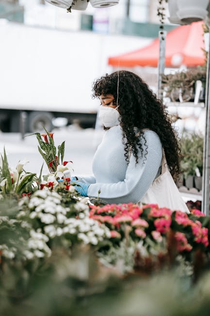 A woman with curly dark hair, wearing a white face mask, a light blue sweater, and blue gloves, is closely inspecting and arranging a bouquet of fresh flowers at an outdoor floral stall. The bouquet f