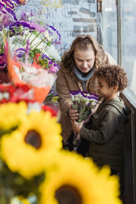 A woman and a young boy stand inside a flower shop near a large window, engaging in a flower delivery or gifting moment. The woman, with light brown curly hair, is gently leaning toward the boy, smili