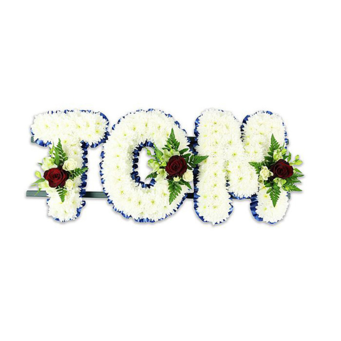 A striking, lovingly arranged name tribute that reads TOM, fashioned from countless small white chrysanthemum blooms that have been placed so closely they form plush, tactile letters. The chrysanthemums' layered petals give a soft, cushion-like volume to each character, while a vivid royal blue ribbon crisply outlines the edges, lending formal definition and a measured sense of grace. Within each letter sits a thoughtfully composed spray: at the heart of every cluster a single deep red rose commands attention-a timeless emblem of affection-surrounded by petite creamy-white spray roses and tight buds that brighten the composition. The floral accents are interlaced with slender fern fronds and varied greenery, their textures ranging from glossy blades to soft, serrated leaves that add shadow and contour. A low dark green or black metal support bar runs behind the letters, visible at the base, hinting at the structural care that keeps the tribute upright during viewings and services. Set against a clean white background and lit with a soft, even light, the arrangement's colours read clearly from afar: the white chrysanthemum field, the royal blue trim and the crimson focal roses combine to create a solemn yet comforting presence. This handcrafted memorial piece is well suited for funeral chapels, church halls or graveside services in Malden Rushett and nearby suburbs, offering a personalised, respectful floral farewell crafted by experienced local hands.