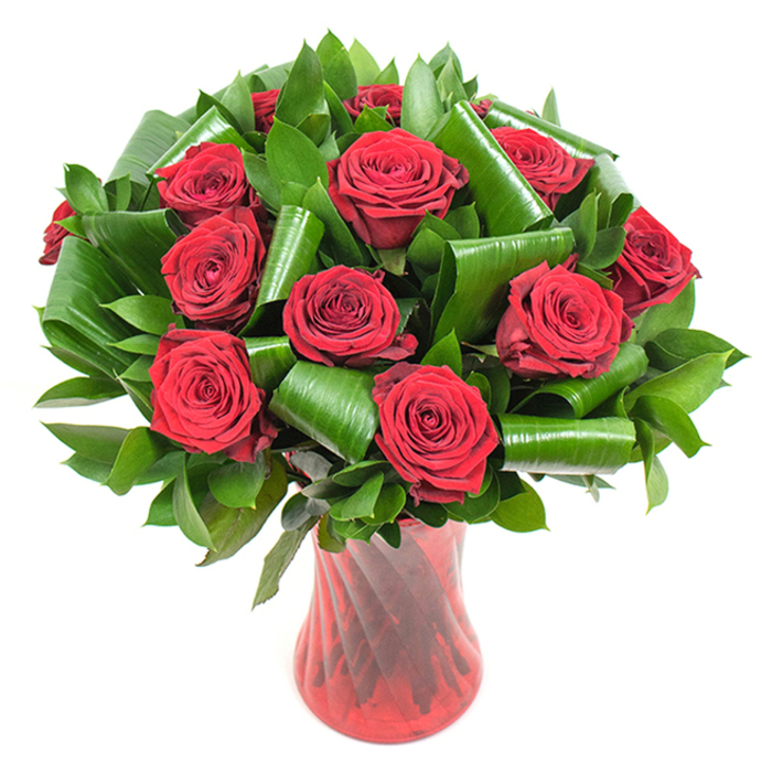 A dozen or more full-blown crimson roses form a dense, rounded bouquet that reads like a study in passion and polish. Each rose is in full bloom, petals tightly spiraled and layered to create that familiar velvet depth where light pools in the folds. Around the rose heads a lush architecture of dark green foliage provides volume and contrast: smaller, glossy leaves create a verdant bed while broader, Aspidistra-like leaves have been skillfully curled into tubular and spiral shapes, lending an almost architectural rhythm to the composition. The bouquet is housed in a tall, translucent ruby-red glass vase whose ribbed, slightly twisted surface throws vertical highlights that echo the stems and folded leaves; the vase's narrow waist and stable base give the whole display an upright, refined silhouette. The image is set against a crisp white backdrop with soft, even illumination that allows the deep reds and emerald greens to pop with clarity. Close your eyes and you might sense a classic rose perfume-soft, familiar, and quietly romantic. As an experienced floral designer in the Malden Rushett area, I can see this piece working beautifully as an anniversary centrepiece or a deluxe Valentine's delivery; it's the kind of arrangement we craft at Flower Delivery Malden Rushett for clients who want traditional romance expressed with contemporary leafwork and clean presentation. The overall impression is luxurious, considered, and heartfelt.