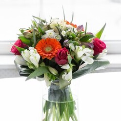 Gorgeous and full of life, this masterful bouquet is a celebration of colour and expert design-a familiar sight crafted with care by local florists in the heart of Malden Rushett. A circle of creamy, softly ruffled gerberas forms a gentle halo, each petal coaxing light towards bright orange centers that spark with cheerful energy. Tucked among these cheerful blooms, rich fuchsia roses lend depth and an air of sophistication, while loose sprays of ivory-hued alstroemeria and petite white filler blossoms whisper of quiet elegance. Verdant foliage spills generously from within, with broad aspidistra leaves skillfully curved to protect and shape the bouquet's full profile-calling to mind the lush hedgerows and meadows that border the Rushett countryside. Sunlight from a broad window floods the arrangement, casting subtle highlights and shadow that accentuate every nuance. The bouquet rests serenely in a tall, clear vase, its shimmering surface echoing the freshness and purity of recent rain on Church Lane. In the air lingers a clean, invigorating scent: lilies, roses, and cut greenery entwined in a fragrant embrace. This floral design feels right for occasions celebrating new chapters-perhaps a move to a new home, a weekend brunch, or an unexpected gesture for neighbours-where each flower is chosen to bring optimism, beauty, and heartfelt care to Malden Rushett's cherished community.