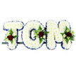 Displayed horizontally on a simple dark frame, this floral name tribute spells TOM in bold, three-dimensional letters, designed as a personalised funeral arrangement for a loved one. Each letter is entirely filled with tightly clustered white chrysanthemums, their petite, rounded petals forming a thick, even surface like a soft floral tapestry. Around the outer edge of each block letter, a royal blue satin ribbon has been neatly folded and secured, tracing the contours of the 'T', 'O', and 'M' with a smooth, gleaming border that offers a striking yet respectful contrast to the clean white blooms. On each letter, a small spray of accent flowers has been positioned with care: deep red roses with velvety petals, small white or cream spray roses, and slim, trumpet-shaped white flowers similar to freesia nestle into a bed of green foliage and fern fronds. These clusters sit, for example, on the upper left of the 'T', along the curve of the 'O', and at the upper right of the 'M', giving balance and rhythm to the design. The tribute is photographed against a plain white background so every petal, ribbon edge, and leaf is clearly visible. It is the kind of bespoke floral piece often chosen by families in Malden Rushett to rest alongside the coffin or at the front of a chapel, holding Tom's name in flowers as a final gesture of remembrance, symbolising purity, love, and peace through its white, blue, and red colour palette.