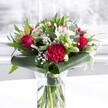 A striking red and white bouquet stands proudly in a clear cylindrical glass vase, its neat green stems visible through the fresh water, as if just arranged on a light-filled table in a Malden Rushett home. Deep red roses, their velvety petals beginning to unfurl, cluster at the heart of the display, each bloom echoing classic romance and heartfelt passion. At the front, a large crimson carnation, densely ruffled and intricately layered, provides a rich textural focal point that draws the eye. Around these bolder flowers, white and creamy blossoms create a gentle contrast: elegant white Alstroemeria with fine speckles on their inner petals, dainty daisies or chrysanthemums, and several closed white lily buds that hint at fragrant blooms yet to open, like promises of tomorrow. Sprigs of delicate white gypsophila, or baby's breath, float throughout the design like tiny stars, adding airiness and soft movement. The lush greenery is just as considered as the flowers, with broad, glossy dark green tropical leaves carefully curled and wrapped around the base, forming an elegant collar that frames the arrangement. Eucalyptus-like foliage and pointed green leaves layer in different tones of green, from deep forest to softer sage, giving depth and structure. The bouquet sits on a pristine white surface with a bright, blurred backdrop that suggests a sunlit window overlooking the quiet streets of Malden Rushett, perhaps not far from the open spaces leading towards Chessington. The even, natural lighting highlights each petal and leaf, creating an impression of freshness, celebration, and warmth. This arrangement feels perfect for an anniversary, a birthday in a local Malden Rushett cottage, or as a refined gift to brighten a neighbour's kitchen table, carrying an atmosphere of joy, admiration, and sincere affection.