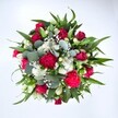 A richly detailed top-down photograph of a circular, hand-tied bouquet shows a generous cluster of deep crimson roses and ruffled ruby carnations, all set against a pristine white background that makes every colour sing. The velvety red rose heads, some still in tight bud and others gently unfurling, sit at the heart of the design and give the arrangement a romantic, almost timeless feel. Around them, full, frilled carnations in shades of fuchsia and cherry red add volume and texture, their petals creating soft, rippling layers. Between the bold reds, small white spray chrysanthemums and other daisy-like blooms punctuate the bouquet with crisp, bright centres, offering a clean contrast that feels fresh and uplifting. Tiny stars of white gypsophila, or baby's breath, are threaded throughout, creating a light, misty veil that softens the transitions from one flower to the next. Lush greenery frames the entire posy: strong, pointed dark green leaves, likely from lilies or alstroemeria, reach outward to define the shape, while rounded, silvery eucalyptus leaves nestle closer in, lending a contemporary softness and suggesting a cool, soothing fragrance. The bouquet appears dense, carefully balanced, and expertly arranged, with no gaps, as though it has just been finished on a florist's workbench in Malden Rushett, ready for local delivery. The overall impression is one of joyful celebration and heartfelt affection, ideal for Valentine's Day, an anniversary, or a special birthday, and perfectly at home brightening a cottage near Chessington Road or a modern flat close to the green edges of Epsom Common. The image captures a sense of abundance and care, as if this bouquet has been crafted to make someone in Malden Rushett feel deeply cherished and remembered.