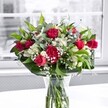 A lush red and white bouquet sits proudly in a classic clear glass vase on a bright white tabletop, bathed in soft daylight from a large, blurred window behind. At the heart of the arrangement, deep crimson roses with velvety petals open generously, while a few tighter buds promise more beauty to come, their rich colour suggesting romance and warm affection. Nestled beside them are full, ruffled red carnations, their layered petals adding volume and a traditional charm that feels perfect for heartfelt occasions in Malden Rushett, from quiet family dinners to special anniversaries. Around these bold reds, clusters of white alstroemeria stretch gracefully, some still in green-tipped bud, others partially open to reveal their slender, trumpet-like blooms, lending a gentle brightness and elegance. Tiny white baby's breath threads through the bouquet like a soft mist, creating a cloudlike halo that lightens the composition and draws the eye back to the stronger colours. A collar of broad, glossy green leaves cradles the flowers, while finer, lighter foliage weaves between the stems, adding depth, movement, and a sense of freshness, as if the bouquet had just been gathered from a garden near the open spaces of Chessington or Epsom. Through the water in the vase, the cut stems appear crisp and neatly aligned, emphasising the care taken in the design. The neutral, almost grayscale outdoor scene glimpsed through the window recedes into gentle blur, ensuring the vibrant reds, clean whites, and varied greens stand forward with clarity. The whole display feels like a polished yet welcoming gift-ideal for saying thank you, celebrating love, or simply brightening a Malden Rushett home with the quiet joy of fresh flowers.