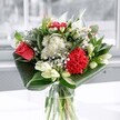 A contemporary glass-vase arrangement stands in soft daylight, its colours and textures unfolding like a quiet celebration in a Malden Rushett sitting room. At the centre, a coral-red rose opens in velvety spirals, its plush petals echoing the deep crimson of a ruffled carnation nearby. These rich reds feel like a steady heartbeat, gently framed by clusters of daisy-like white chrysanthemums and tiny button blooms that bring an airy, almost cloud-soft lightness to the design. Around and between them, creamy white lilies rest mostly in bud, their long, sleek forms adding poise and anticipation, as if the bouquet is caught at the very beginning of its fullest beauty. Broad, glossy emerald leaves sweep in confident arcs around the base, carefully folded and layered by the florist to cradle the stems and give the composition a clean, architectural silhouette. The clear glass vase reveals the stems gathered neatly together, water glinting faintly where the window light touches it. The atmosphere is crisp yet tender, like a fresh morning stroll along the lanes near Chessington World of Adventures, with a hint of leafy fragrance and gentle sweetness in the air. This is the kind of bouquet you might place on a kitchen table in a Malden Rushett cottage to mark a birthday, an anniversary, or simply to lift a grey day, its balance of passionate reds and peaceful whites offering both energy and calm in one thoughtfully arranged piece.