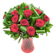 A lush, museum-quality bouquet of roughly a dozen deep crimson roses fills the frame, each bloom opened fully to reveal tightly spiraled, velvety petals that catch the light like polished silk. The intense, wine-dark red of each head is offset by a generous collar of glossy dark green foliage - smaller Ruscus- or Salal-like leaves that form a dense backdrop and broader, emerald Aspidistra-style leaves that have been artfully curled and rolled into architectural spirals. Those glossy loops lend a contemporary silhouette to the classic rose, their satin surfaces reflecting the soft, even studio light and creating gentle highlights along each curve. The stems rest in a tall, slender ruby-red glass vase whose translucent body shows subtle vertical ribbing and a faint twisted texture; the vase's narrow waist and flared lip create an elegant posture that echoes the vertical lines of the leaves. Set against a clean white background, the reds read as especially saturated and the greens appear luminous, isolating the arrangement so the eye travels from the petal edges inward to the heart of each bloom. If you imagine the scent, it is a quietly heady, classic rose perfume - familiar and comforting. This composition reads as a refined romantic statement: the kind of bouquet chosen for an anniversary to be delivered locally around Malden Rushett, or to mark a special dinner at home. The overall mood is one of quiet luxury and thoughtful craftsmanship, where traditional symbols of passion meet considered, modern floral architecture.