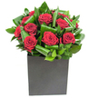 A confident, modern bouquet centred on deep crimson roses that read as both romantic and refined. Each rose opens in velvety spirals, petals layered with a plush, tactile depth that seems to catch the light like satin. Around those blooms, broad glossy leaves have been carefully curled and overlapped to create a rhythmic pattern of green-like waves of emerald that both cradle and lift the roses. The flowers sit in a matte black square box that anchors the composition with a sculptural, urban edge; its flat surface absorbs light so the crimson tops feel to glow against a darker base. Lighting here is clean and steady, bringing out the velvet sheen of the petals and the glassy highlights on each leaf, suggesting an interior setting-a city apartment or a reception desk-where details can be admired close up. There is a faint scent where rose and fresh foliage meet: classic, warm rose softened by green, almost tropical notes from the folded leaves. As a local florist I imagine this arrangement placed on a small dining table after a walk through nearby Chessington lanes or delivered to mark a thoughtful anniversary in New Malden; it reads as a deliberate declaration rather than a casual gift. Every curve and fold in the design has been chosen for balance and movement, the kind of composition that looks as at home on a sleek office counter as it does brightening a loved one's hallway on a rainy Surrey afternoon. It's an elegant, tactile expression of affection that feels carefully considered and immediately present.